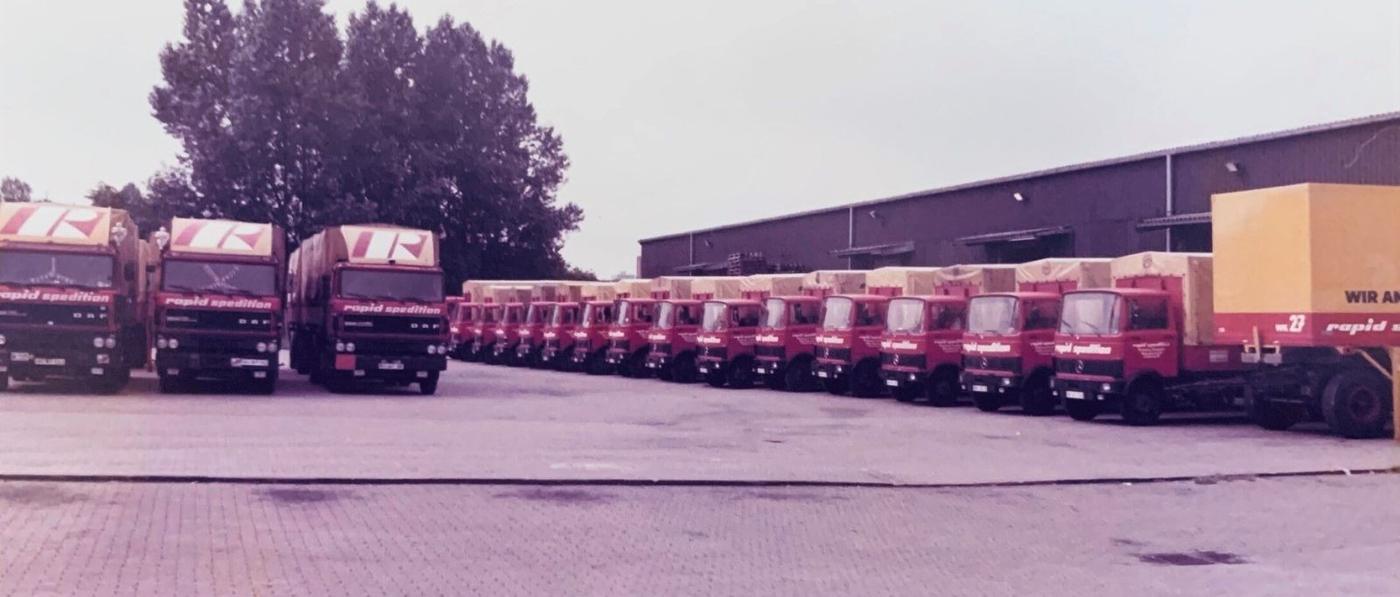 Lange Reihe roter Rapid-Lkw mit gelben Planen vor der Lagerhalle, Ende der 1980er Jahre